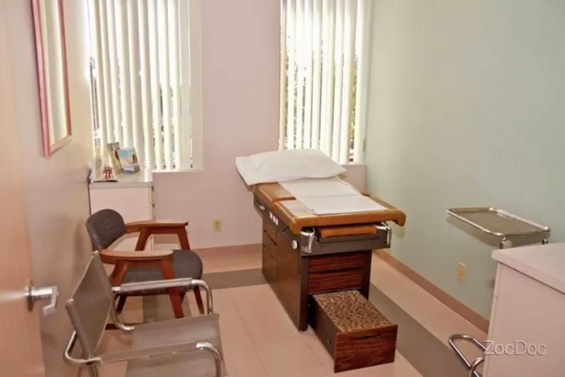 An exam room at the office: a padded exam table with a step stool, two chairs and a counter, under two curtained windows