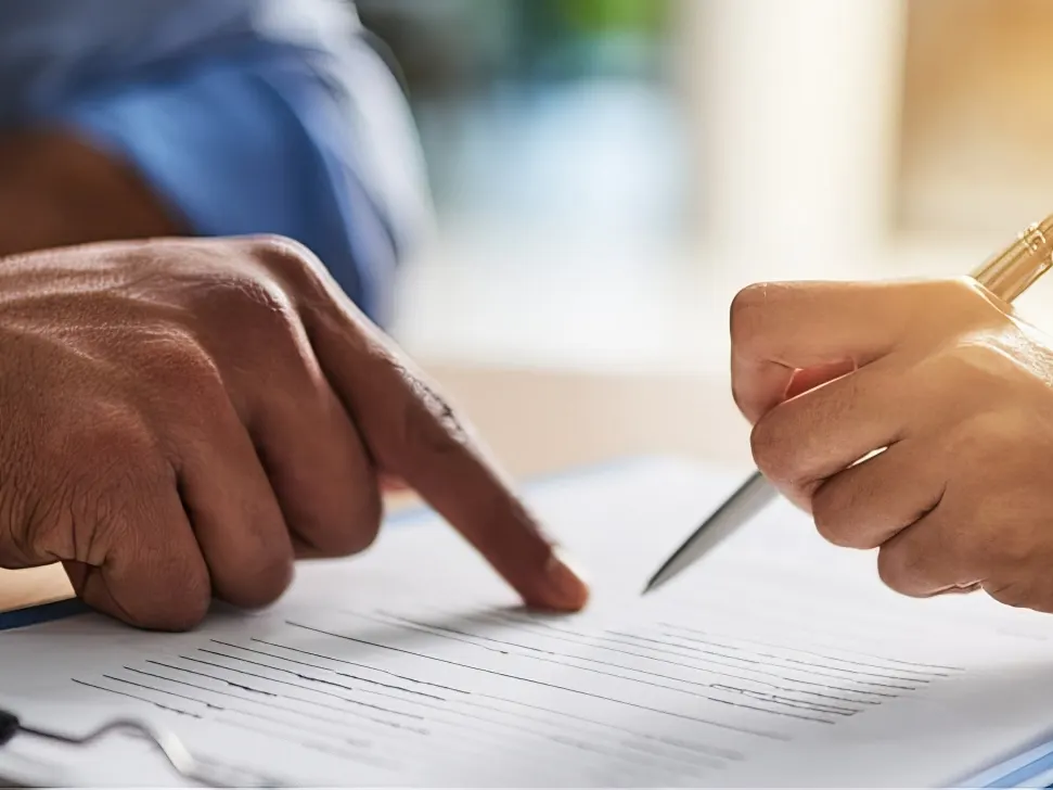 Stock photograph of models: two people at a table, one pointing at a printed form on a clipboard while the other holds a pen