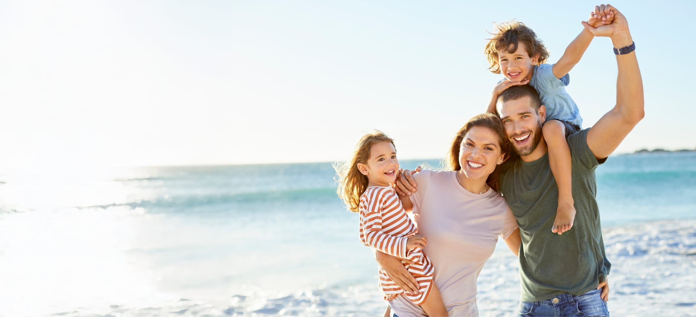 Stock photograph of models: a smiling family on a sunny beach, the father carrying a young boy on his shoulders and the mother holding a little girl