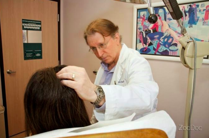 A doctor in a white coat and glasses parting the hair of a seated person with one hand, in an exam room